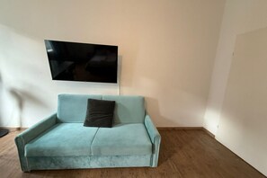 A light blue sofa with a dark cushion, a flat-screen TV mounted on the wall, and a wooden floor. - Beautiful 2BR Apt. With WIFI in a Prime Location (Vienna)
