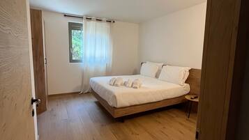 A bedroom with a wooden floor, a bed with white linens, a window with a curtain, and a small bedside table.