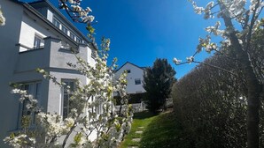 Un edificio residencial con fachada blanca, cerca de piquetes blancos y un jardín bien cuidado con árboles florecientes.