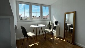 Una habitación con un ventanal grande, una mesa redonda, dos sillas, un mueble bajo y un espejo en la pared.