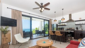 Ein modernes Wohnzimmer mit großem Fernseher, bequemen Sitzmöglichkeiten, einem Deckenventilator und Blick aufs Meer.