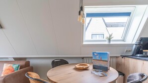 Une pièce chaleureuse avec une table ronde en bois, deux chaises, un canapé avec des coussins, une petite plante en pot et une vue à travers une lucarne.