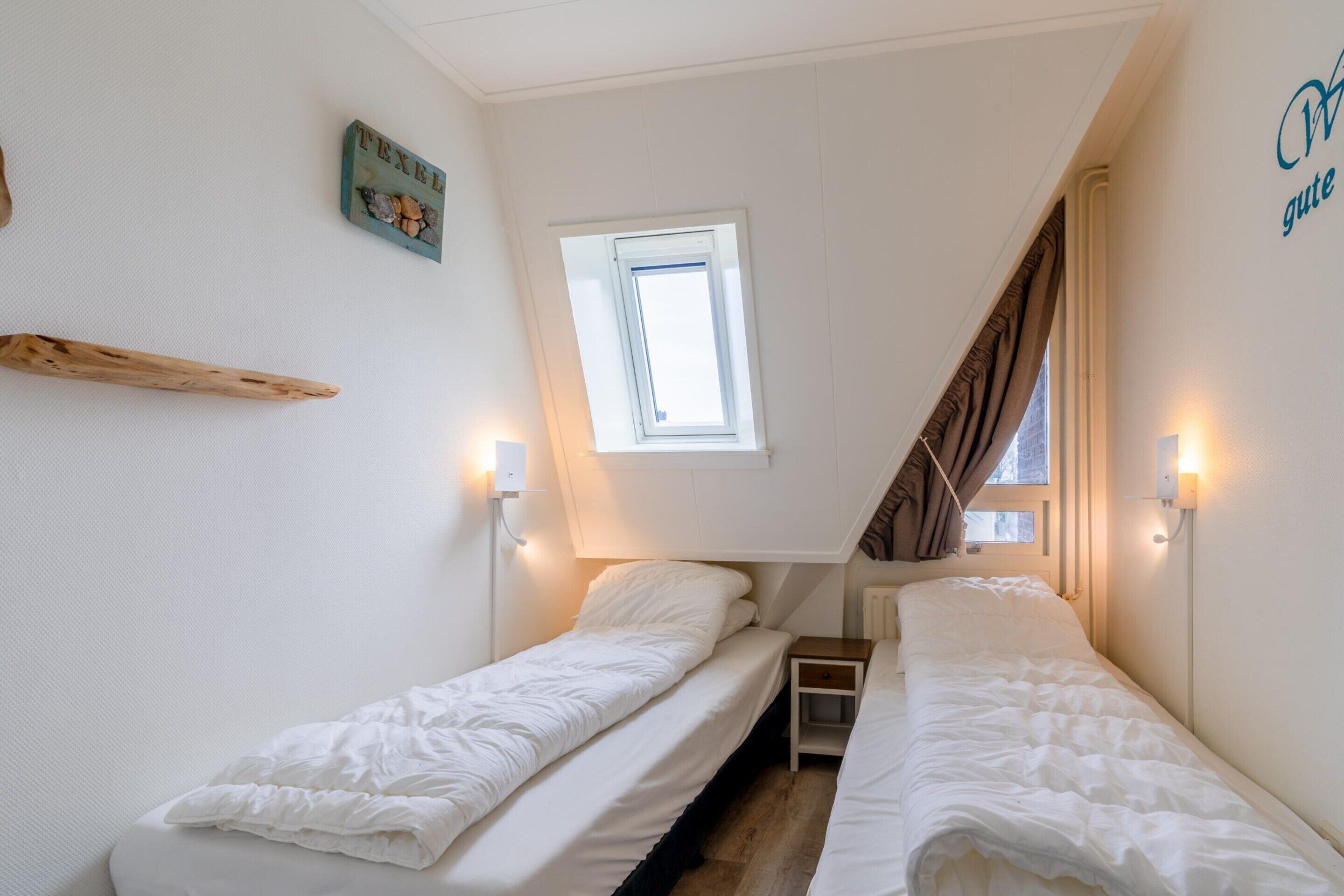 Une petite chambre bien éclairée, avec deux lits simples, une fenêtre et une étagère fixée au mur.