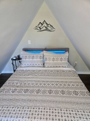 Une chambre à coucher avec un lit, une literie à motifs, une décoration murale représentant une montagne et une table de chevet avec une lampe.
