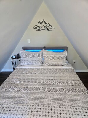 A bedroom with a bed, patterned bedding, a wall-mounted mountain decoration, and a bedside table with a lamp. - Smash Shack | Pickleball Cabin by Red White & Blue Views (Lookout Mountain)