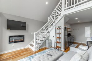 A modern living room with a staircase, a fireplace, a television, and a kitchen area. - Smash Shack | Pickleball Cabin by Red White & Blue Views (Lookout Mountain)