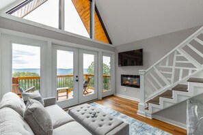 A spacious living room with a grey sofa, a fireplace, and a staircase leading to a balcony with a view of the mountains. - Red White and Blue Views| 4 Cabins + 2 Courts (Lookout Mountain)