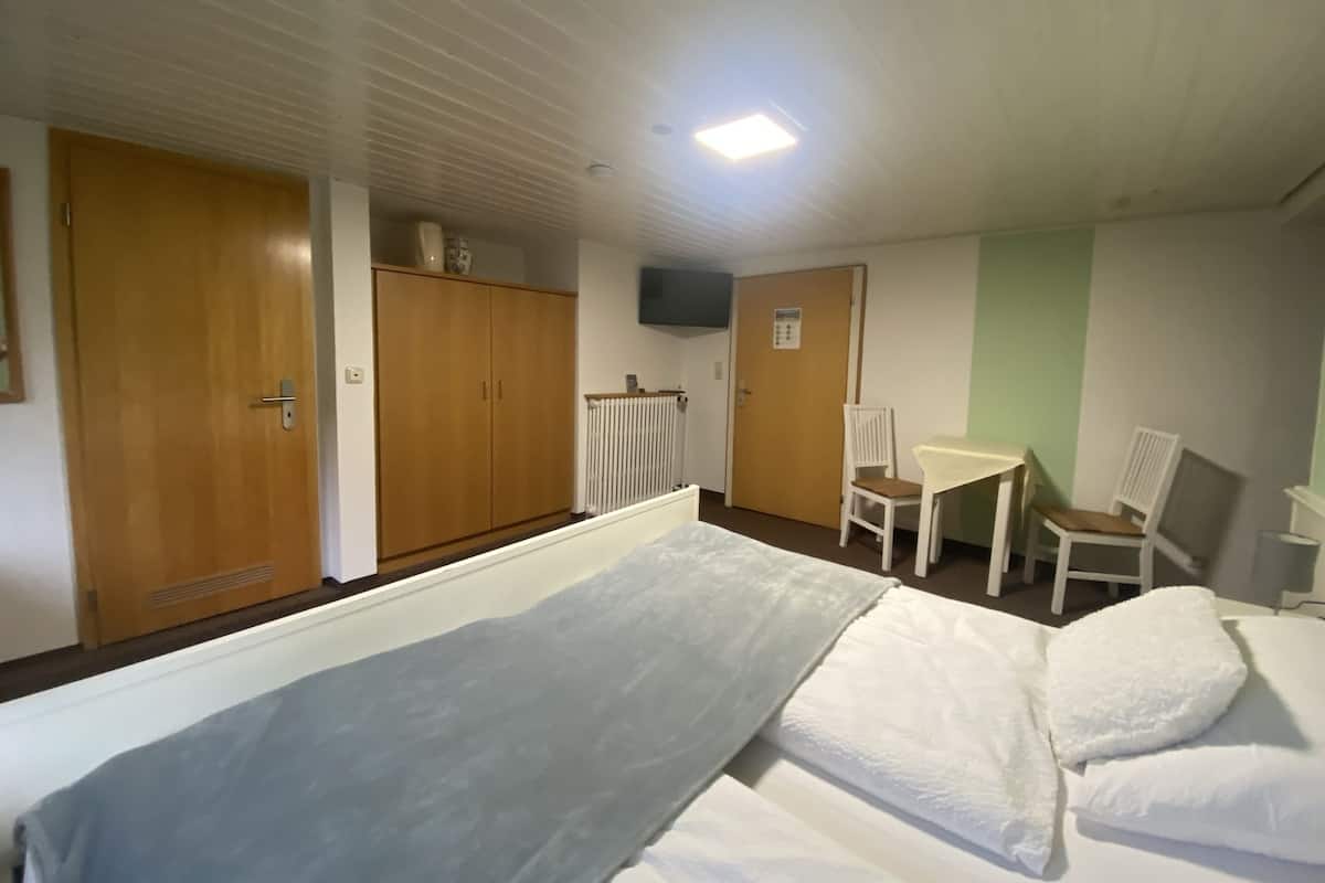Une chambre d’hôtel avec un lit, un bureau, deux chaises, un radiateur et un téléviseur fixé au mur.