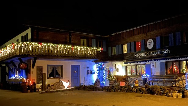 Egy épület, amelyen egy „Nagelfluhhaus Hirsch” feliratú tábla látható, és a balkonját fényekkel díszítették.