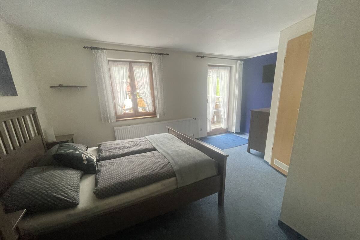 Une chambre à coucher avec un lit, deux fenêtres avec des rideaux, un radiateur et une moquette bleue.