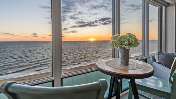 Quarto com vista para o oceano ao pôr do sol, uma mesa redonda com um vaso de flores e duas cadeiras.