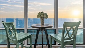 Quarto com duas cadeiras verdes e uma mesa redonda pequena com um vaso de flores, com vista para o pôr do sol sobre o oceano.