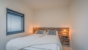 A neatly made bed with a white blanket and pillows, a window with blinds, and a wall with a wooden texture and wall-mounted lights. - Charming 2-person Holiday Home on the Brink in De Koog (De Koog)