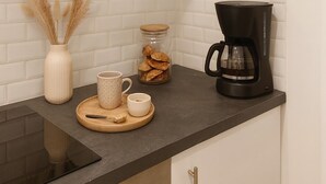 Et køkken med lyse skabe, en sort bordplade, en kaffemaskine, en krukke med småkager, en vase med pampasgræs og en kop.