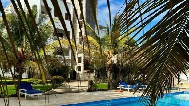 Una piscina con bordo curvo, circondata da palme e da un edificio con facciata bianca sullo sfondo.