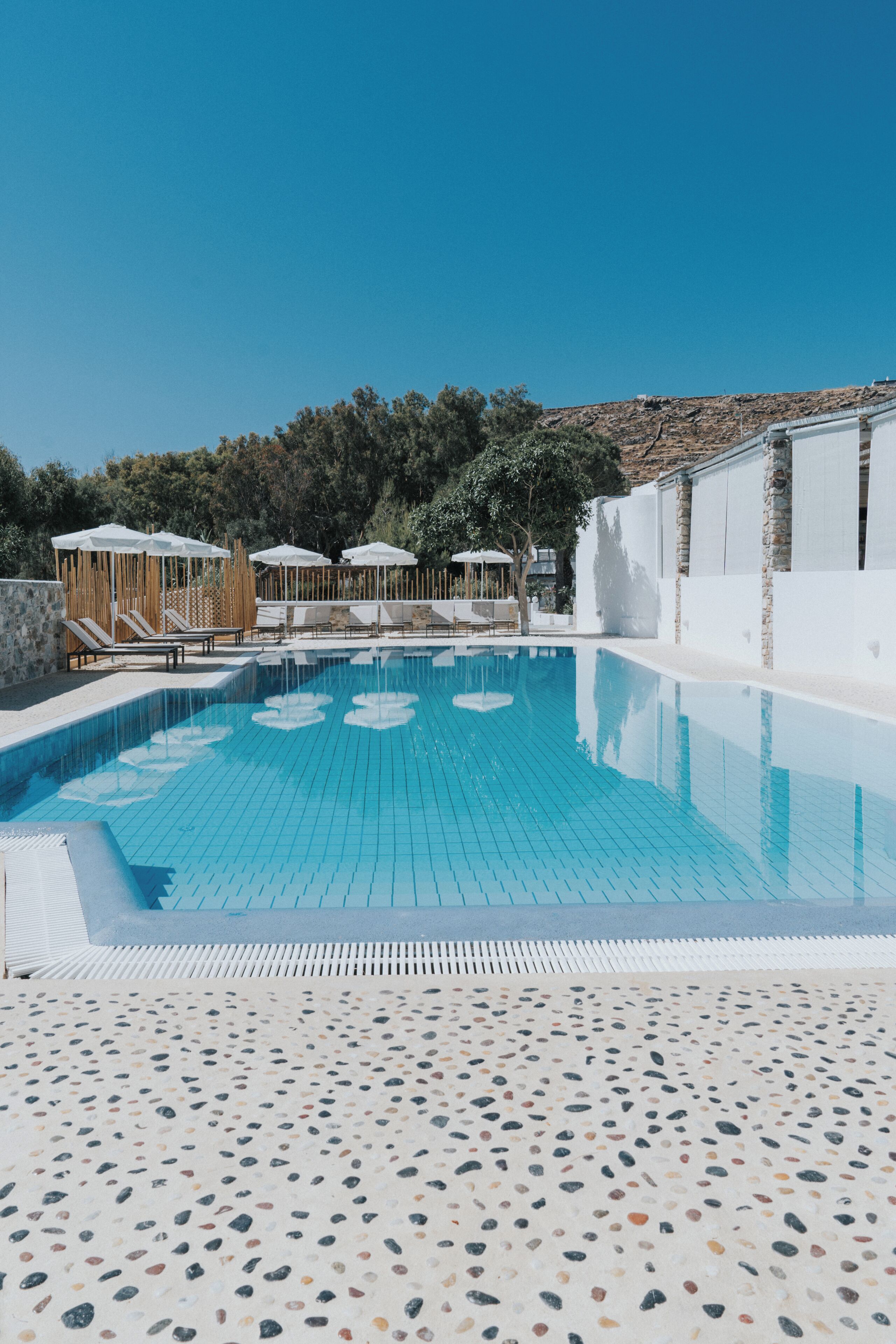 Une piscine d’un bleu éclatant, dotée de chaises longues et de parasols, entourée d’une terrasse au motif de galets et d’un bâtiment blanc agrémenté d’éléments en pierre.