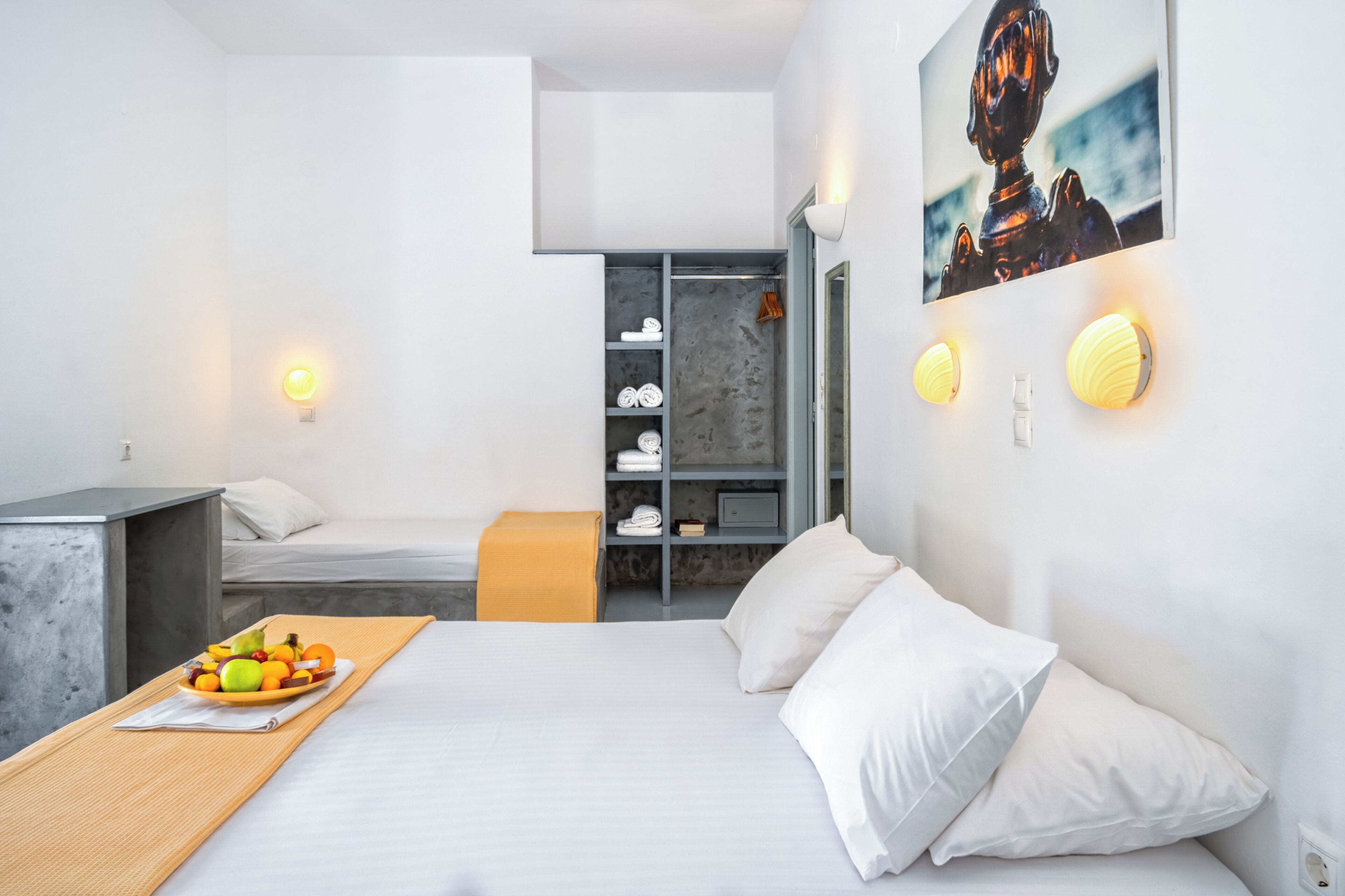 Une chambre d’hôtel moderne, équipée d’un lit, d’un petit bureau, d’un panier de fruits, d’une armoire et d’un tableau au mur.