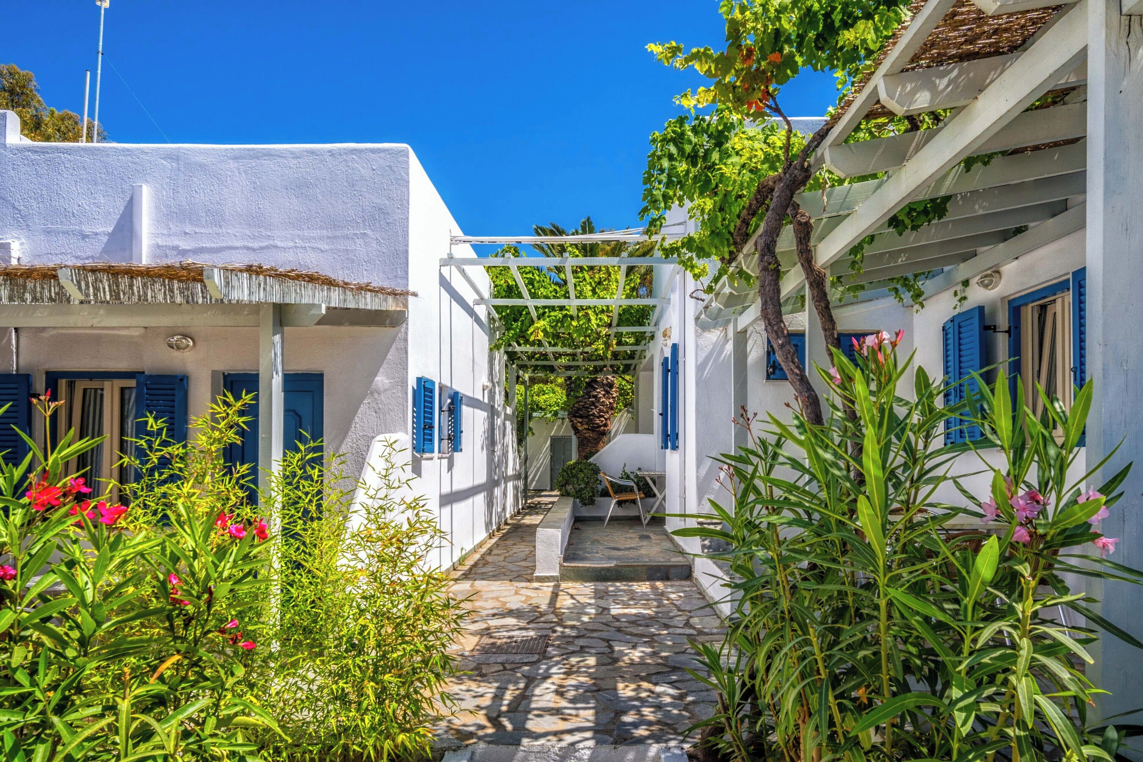A narrow stone pathway flanked by white buildings with blue shutters, leading to a covered area with a table and chairs.