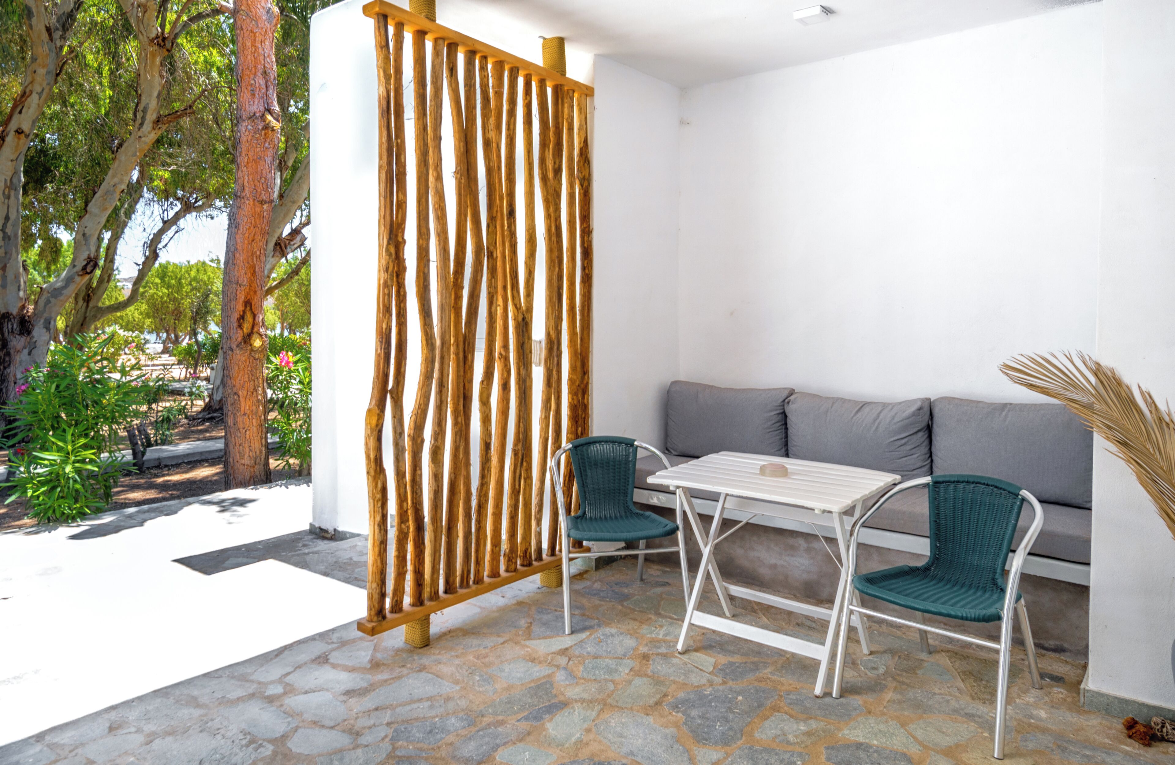 Un patio al aire libre con suelo de piedra, una mesa blanca, dos sillas verdes, un sofá gris y una celosía de madera.