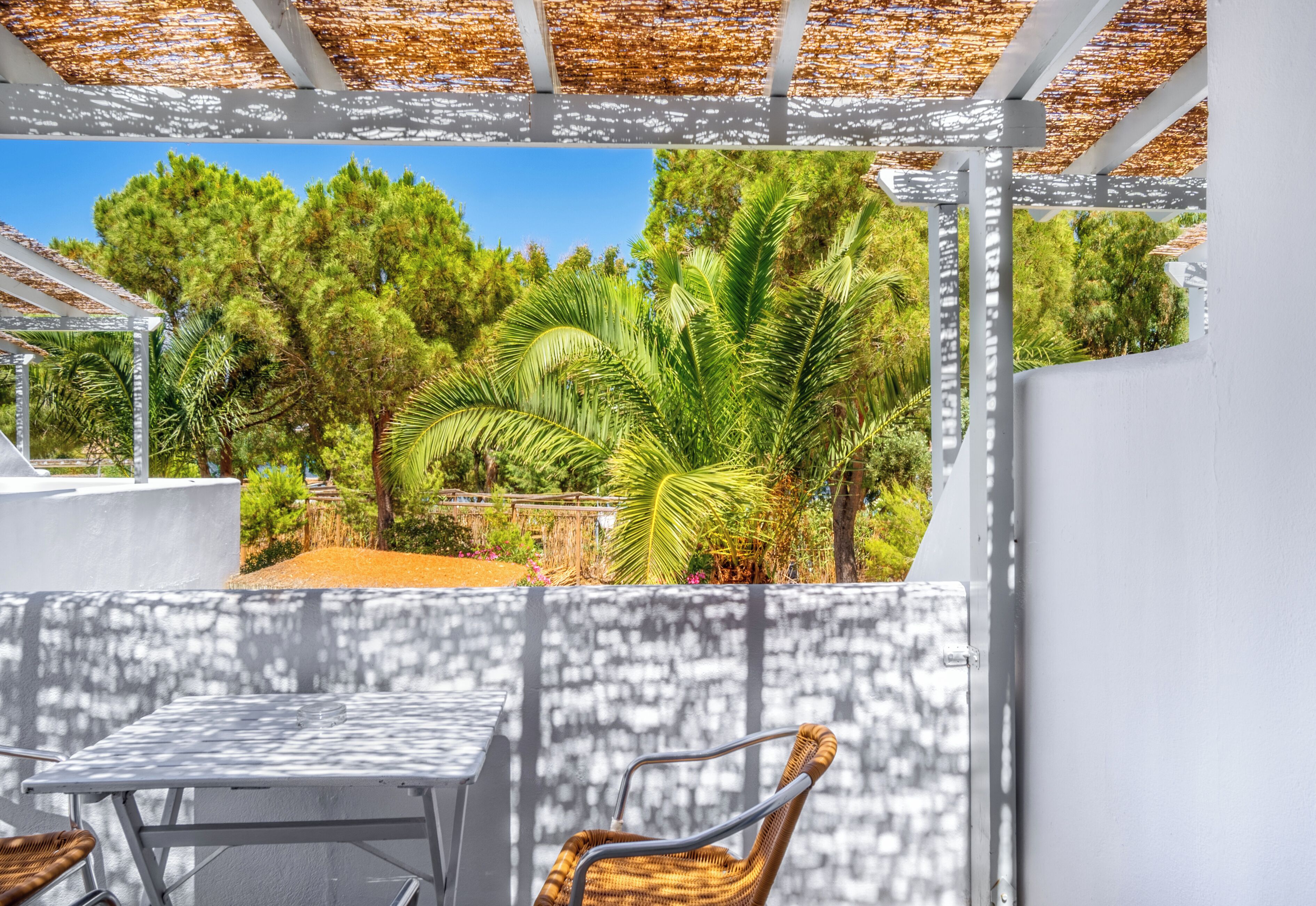 Une terrasse extérieure avec une table blanche et des chaises en osier, entourée d’une végétation luxuriante et surmontée d’une pergola.