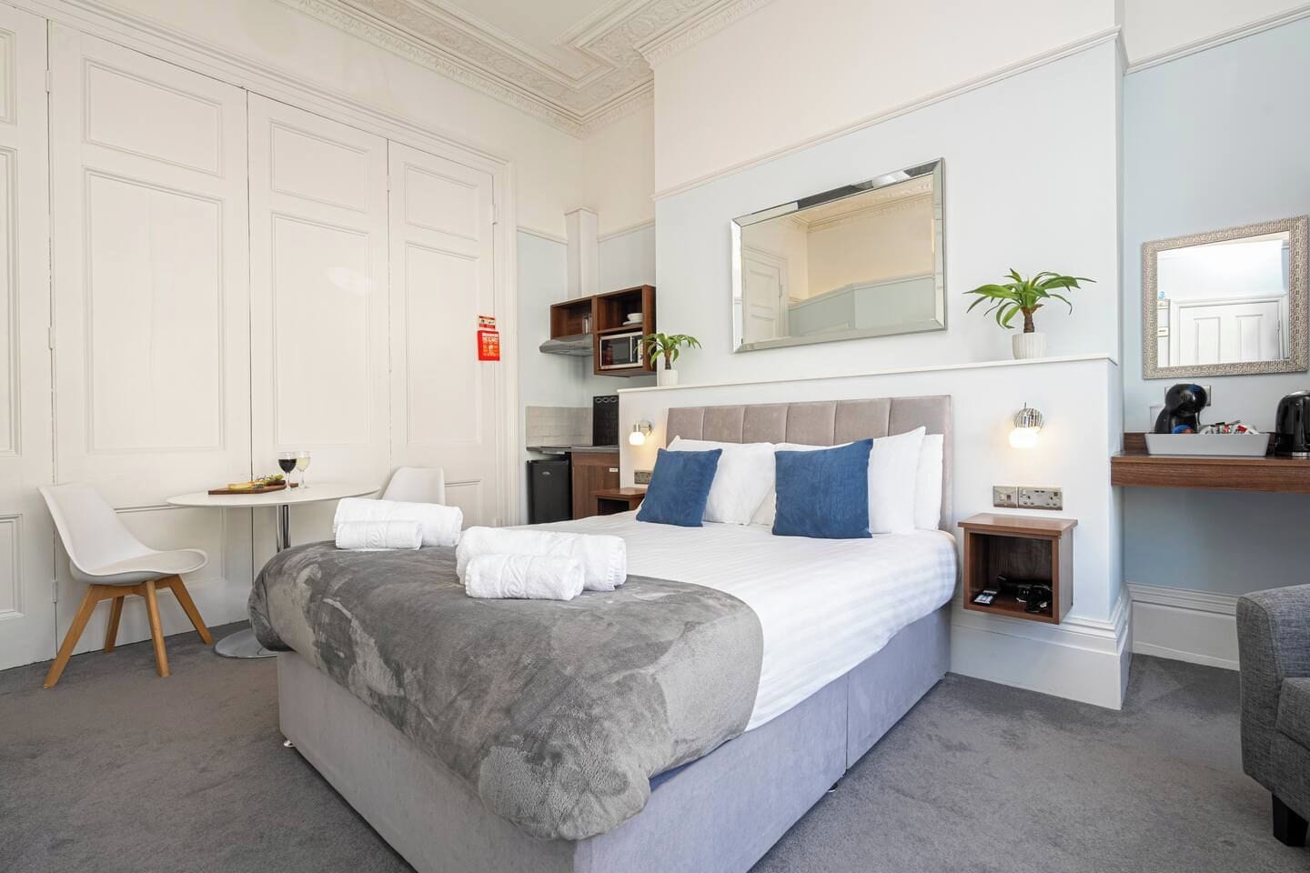 A hotel room with a bed, a desk, a chair, a microwave, a small plant, and a mirror.