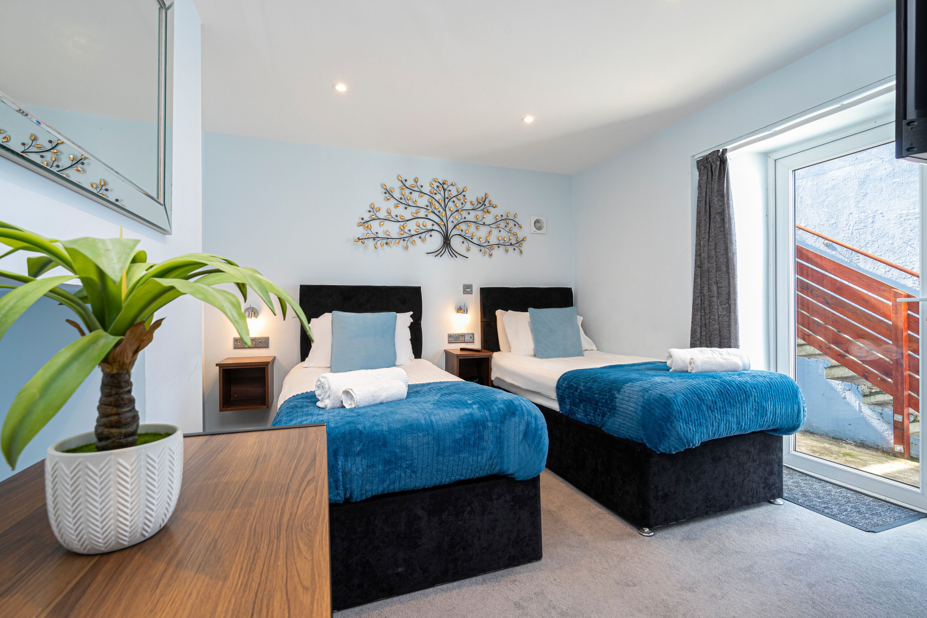 A modern bedroom with two beds, a large potted plant, and a decorative tree wall art.