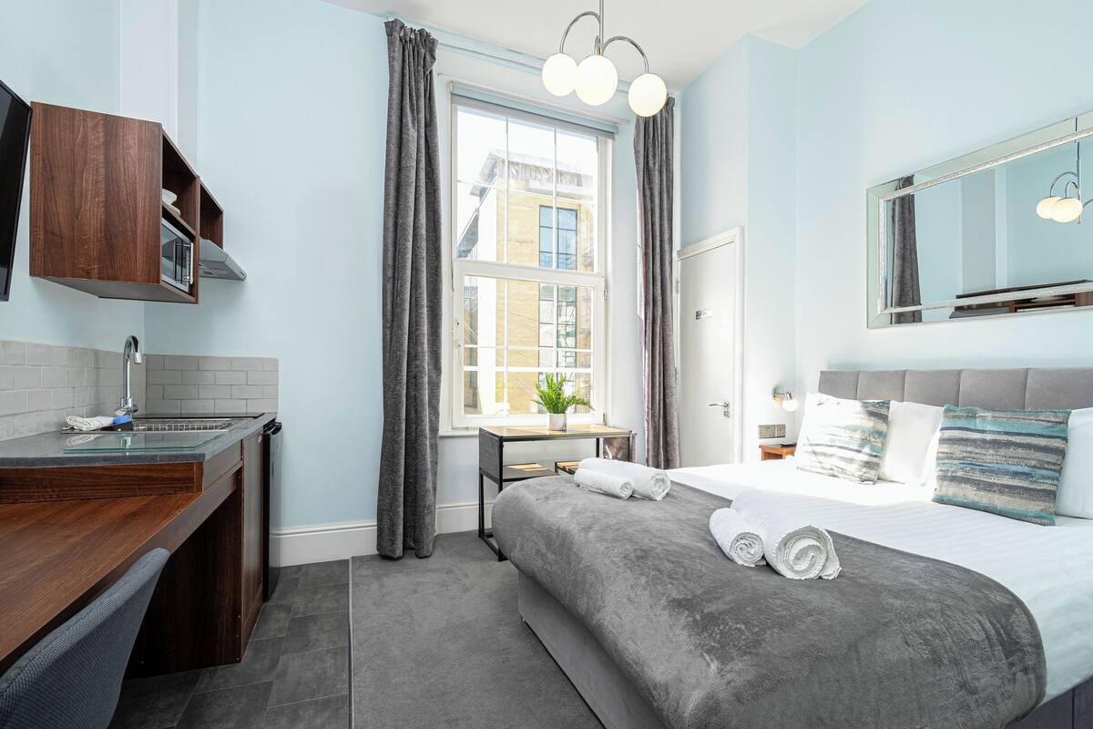 A modern hotel room with a bed, a desk, a window with curtains, a mirror, and a plant.