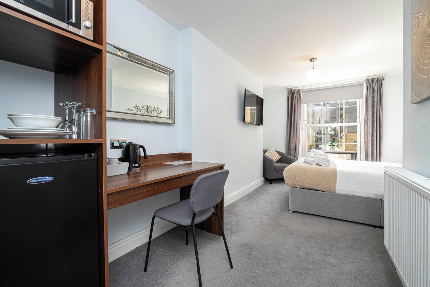 Habitación de hotel compacta con kitchenette, cama, escritorio y ventana con cortinas.