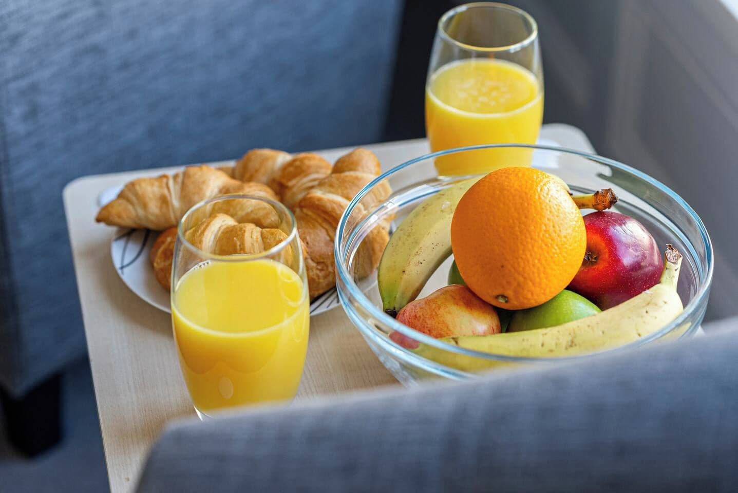 A breakfast tray with croissants, a glass of orange juice, and a bowl of fruit including an orange, apple, and banana.