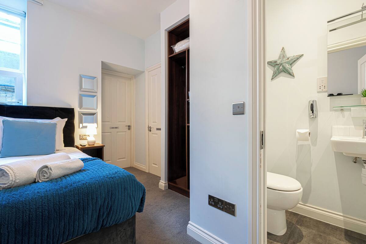 A bedroom with a bed, a nightstand, a window, and a bathroom with a toilet and sink.