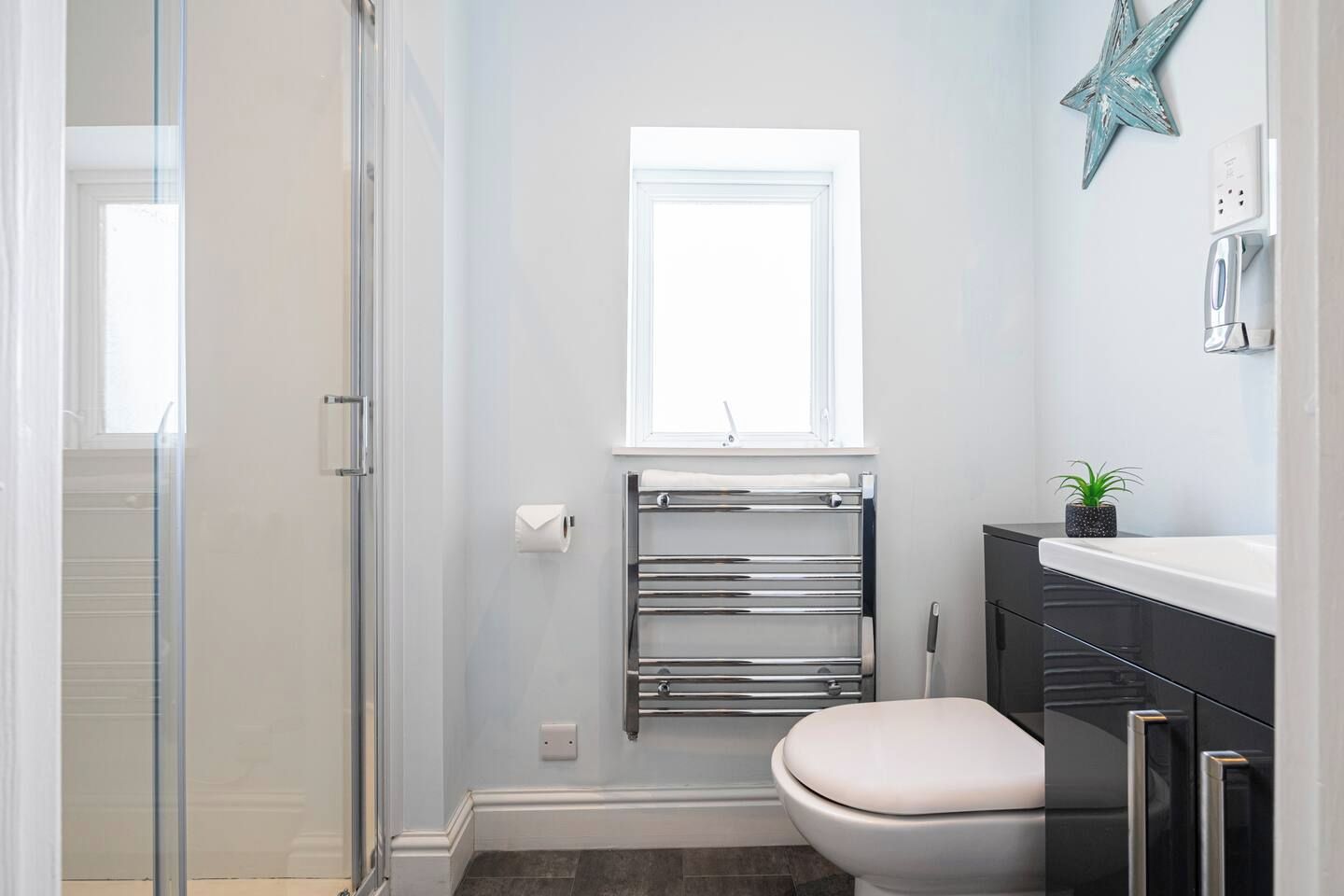 A bathroom with a glass shower enclosure, a toilet, a towel rack, a small window, and a sink with a plant.
