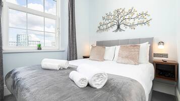 Una cama bien hecha con una manta gris y dos toallas enrolladas, un adorno de árbol en la pared, una mesita de noche con una lámpara y una ventana con vistas a edificios.