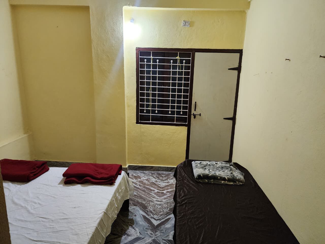 A small, single-room accommodation with two beds, one with a white sheet and the other with a black sheet. There is a window with a metal grid and a door with a black frame.