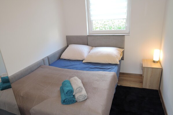A small, neatly made bed with a blue bedspread and two pillows, a bedside table with a lamp, and a window with blinds. - New Holiday Home for 5 People in Wiselka (gmina Wolin)