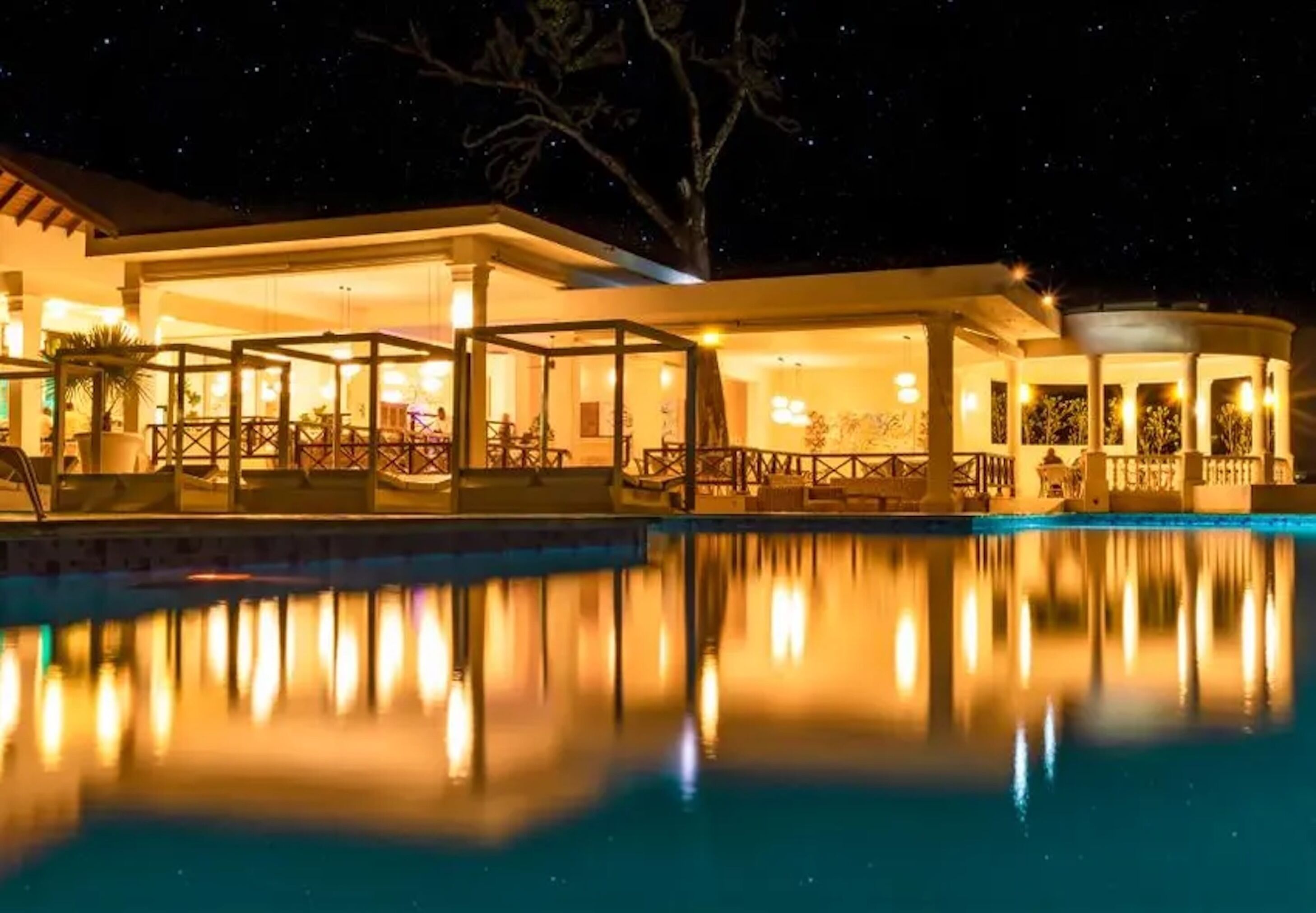 A nighttime view of a hotel with a pool reflecting lights, covered outdoor seating areas, and a large tree in the background.