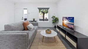 Ein modernes Wohnzimmer mit einem grauen Sofa, einem weißen Couchtisch, einem Flachbildfernseher und einem großen Fenster mit Blick auf Bäume.