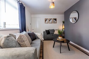 Un salon moderne avec un canapé gris, une table basse et un divan gris foncé. Trois tableaux abstraits encadrés ornent le mur, ainsi qu’un miroir rond et une plante en pot posée sur une table d’appoint.