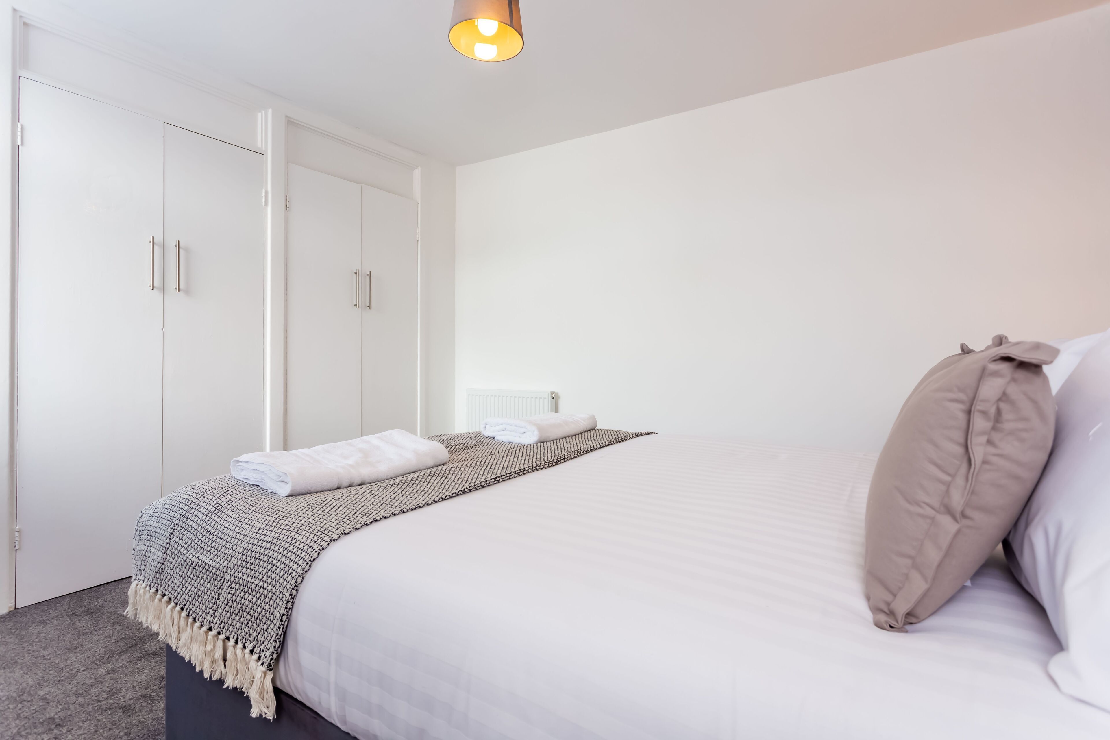 A neatly made bed with a white comforter and a beige pillow, a patterned throw blanket, and a folded towel on the side.