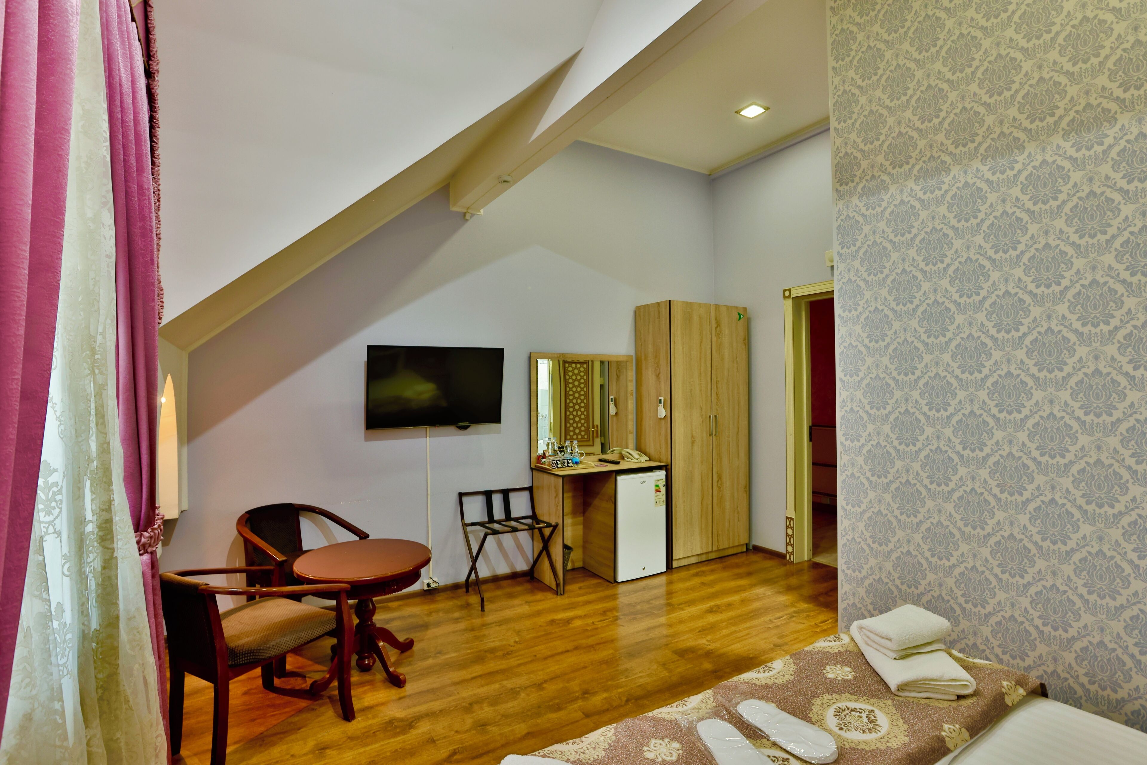 A compact room with a wooden desk, a chair, a small kitchenette, and a TV mounted on the wall.