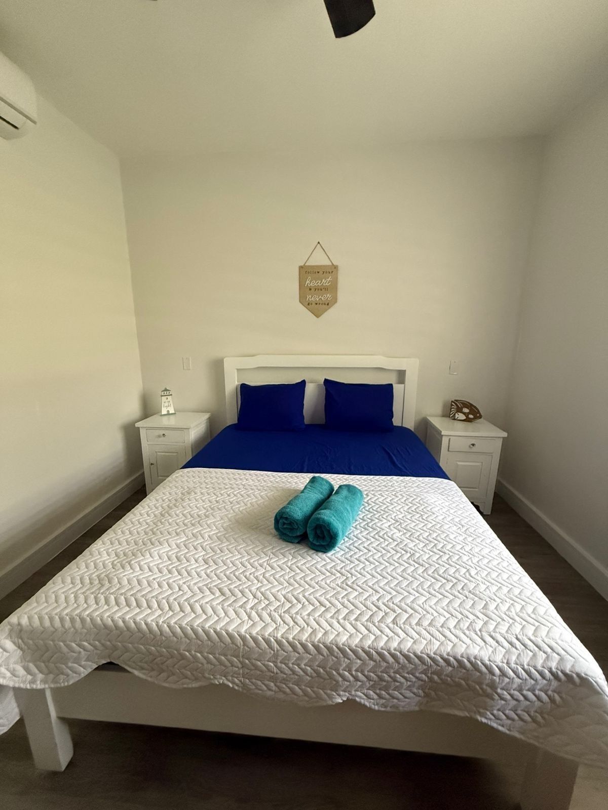 Un lit bien fait, avec une couette blanche et deux serviettes enroulées, une couvre-lit bleue et une petite décoration murale.