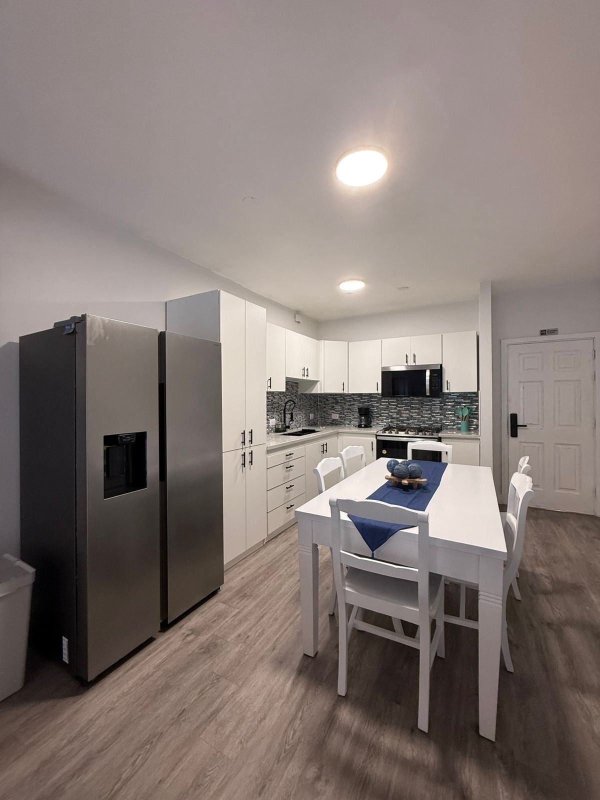 Una cuina moderna amb armaris blancs, un frigorífic d'acer inoxidable, una taula de menjador amb cadires i un forn de microones integrat.