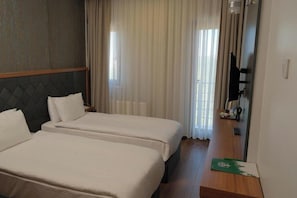 Habitació d'hotel amb dos llits, un panell de capçalera de fusta, un mirall i vista a l'exterior a través de cortines translúcides.