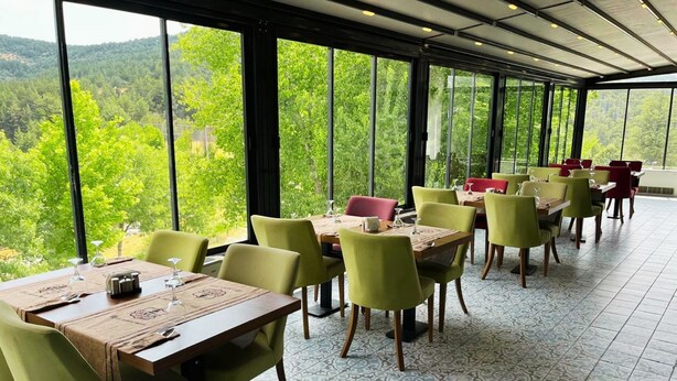 Un restaurant doté de grandes fenêtres donnant sur une forêt. Les tables sont disposées et mises avec des chaises et de la vaisselle.