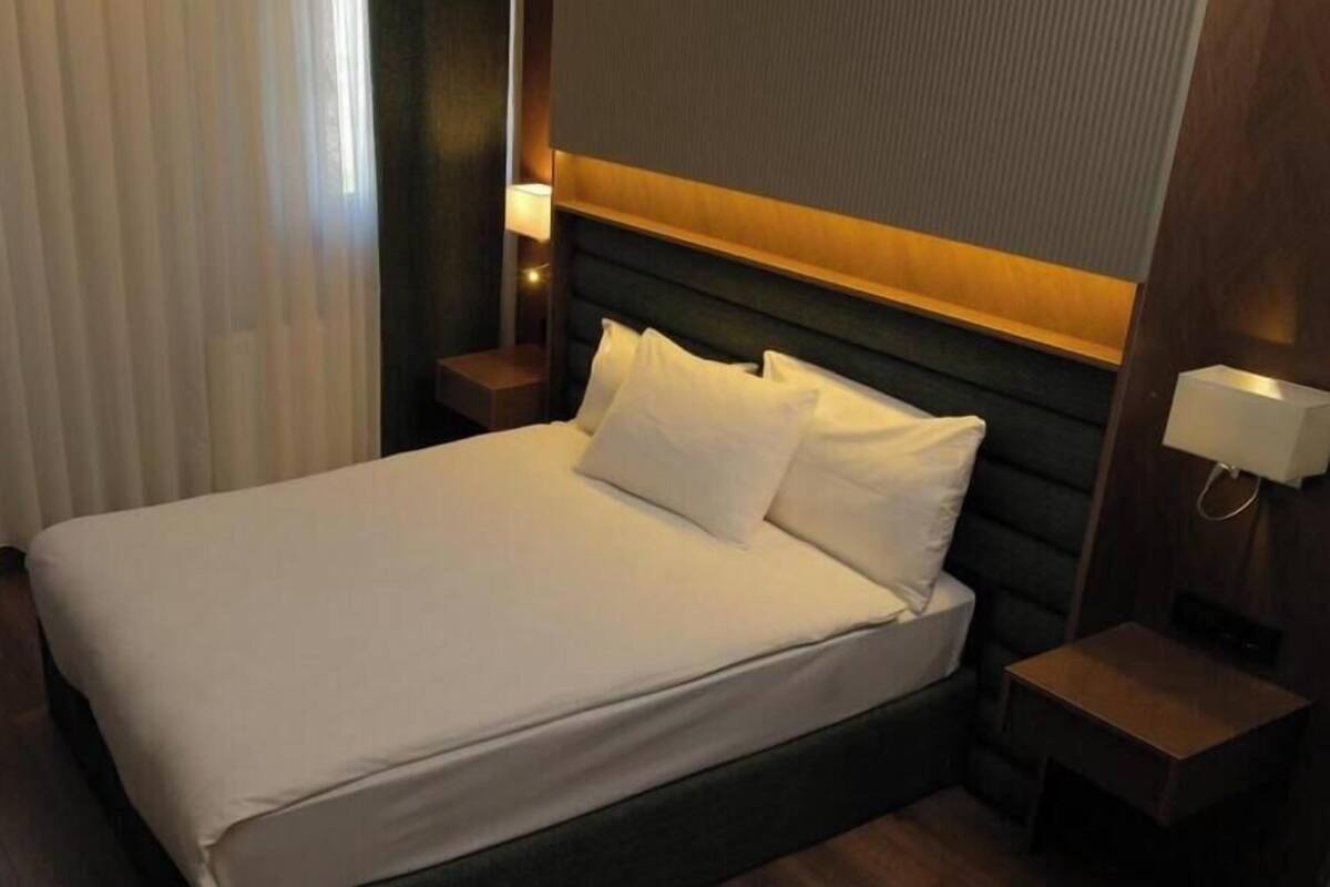 Una habitación de hotel con cama, mesita de noche, lámpara y ventana con cortinas.