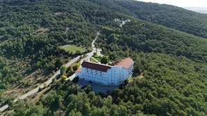 Une vue aérienne d’un grand bâtiment blanc, surmonté d’un toit rouge, entouré d’une forêt dense et bordé d’une route sinueuse.
