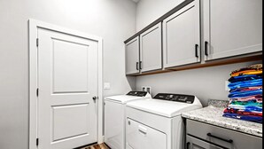 A laundry room with a white door, a washing machine, a dryer, and a countertop with folded clothes. - Game Room • New Construction • 5 Bedrooms, 2 Baths (Mesquite)