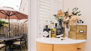 Un'area patio con un tavolo rotondo e sedie, una recinzione in legno e un muro bianco con un armadietto in legno e un vaso di fiori secchi.