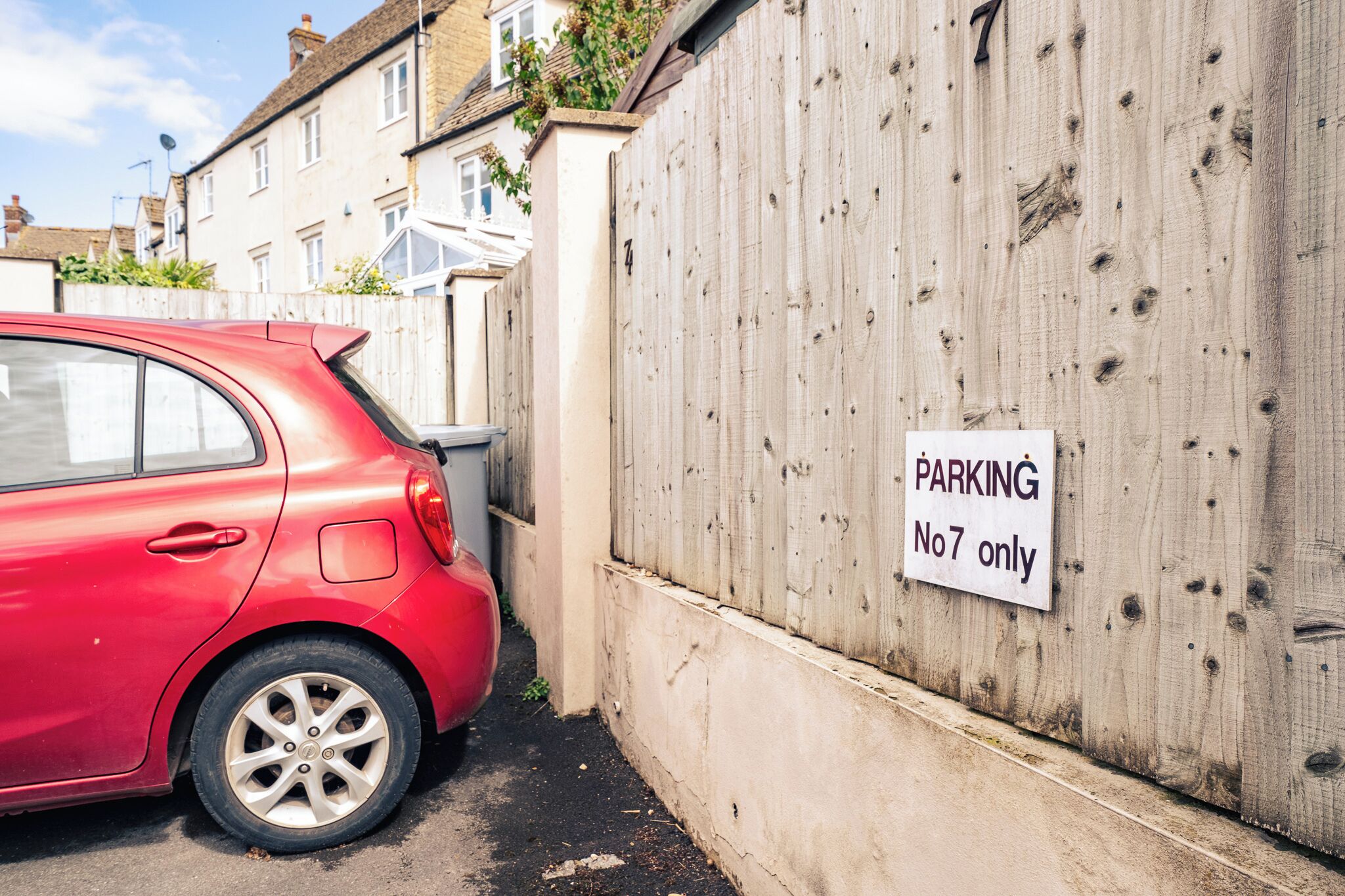 A red car parked next to a wooden fence with a sign that reads "PARKING No 7 only".