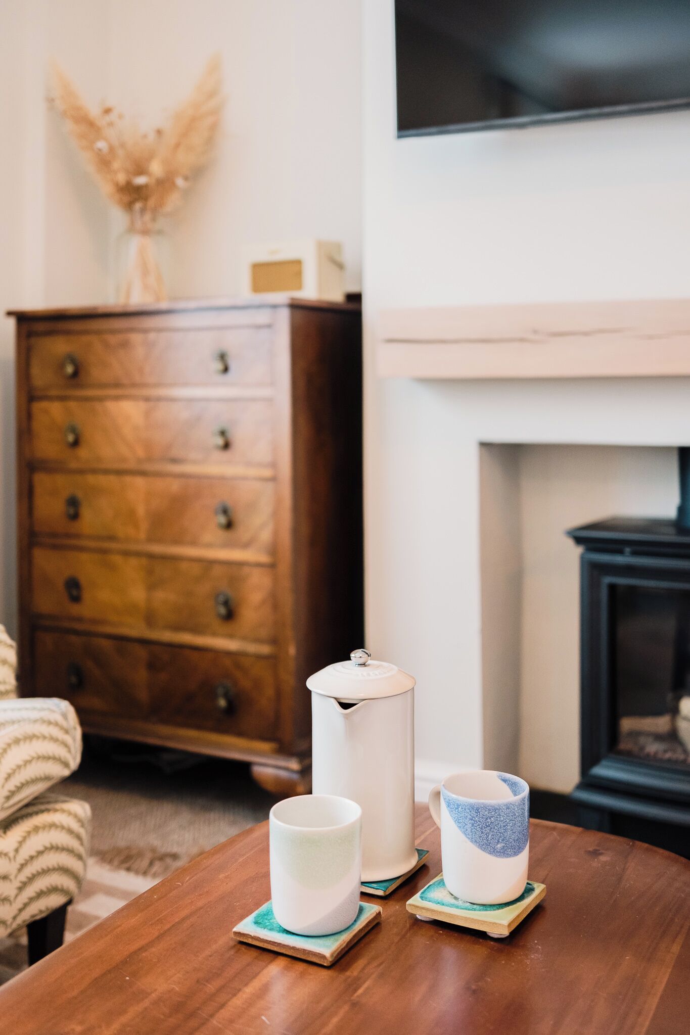 A wooden table with three ceramic mugs on coasters, a vase with dried flowers, and a wooden dresser in a room with a fireplace.