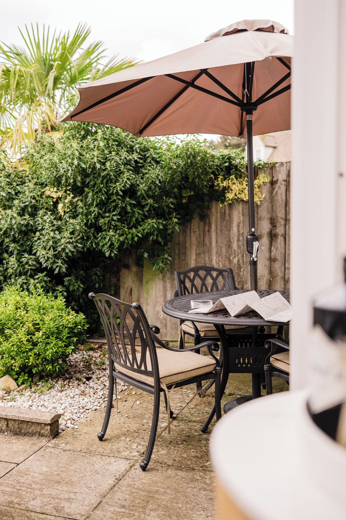 A patio with a table and chairs under a large umbrella, surrounded by greenery and a wooden fence.